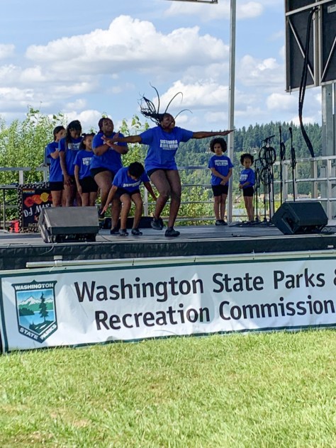 Photo depicting a group of Black-presenting youth performing on a stage with a white banner that reads, "Washington State Parks & Recreation Commission."